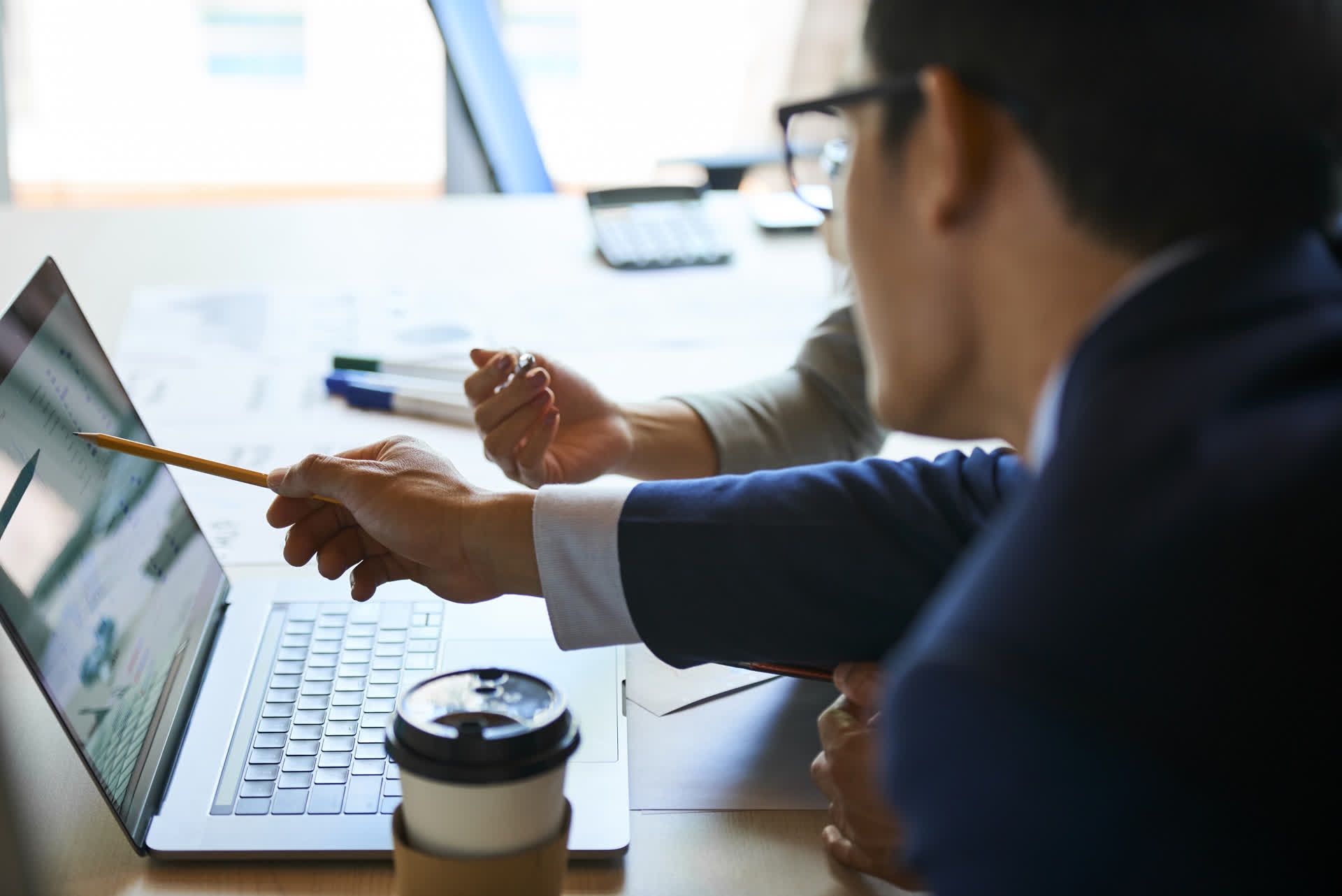 Zwei Personen sitzen an einem Schreibtisch und arbeiten zusammen, wobei eine von ihnen mit einem Bleistift auf einen Laptop-Bildschirm zeigt. Auf dem Tisch liegen Papiere, Stifte und ein Einweg-Kaffeebecher. Der Schwerpunkt liegt auf Zusammenarbeit und Diskussion.