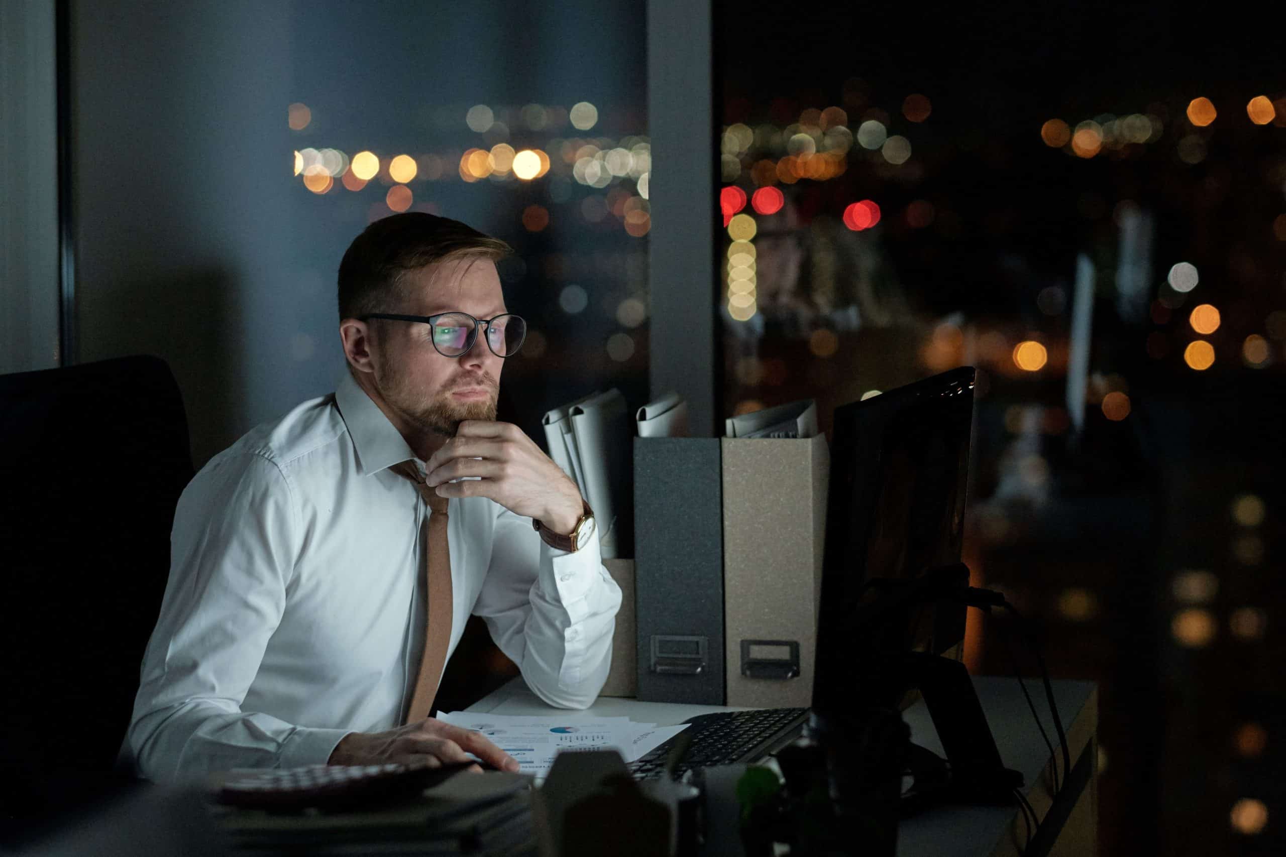 Ein Mann mit weißem Hemd und Krawatte sitzt nachts an einem Schreibtisch in einem schwach beleuchteten Büro und schaut auf einen Computerbildschirm, während die Lichter der Stadt durch das Fenster hinter ihm zu sehen sind.