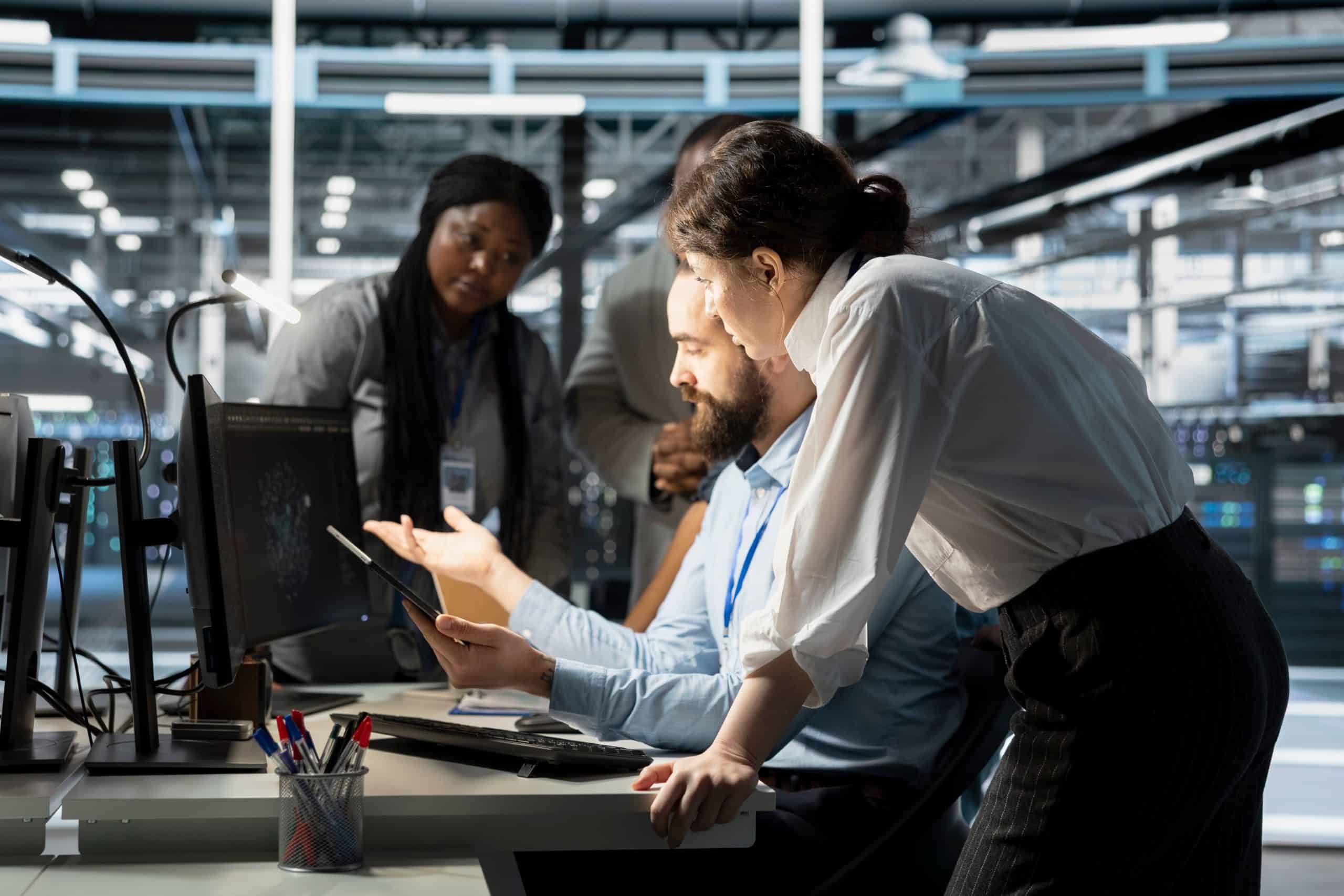 Vier Fachleute arbeiten in einem modernen Büro zusammen. Zwei Personen sitzen an Schreibtischen mit Monitoren, während zwei andere stehen, wobei einer auf ein Tablet zeigt. Die Gruppe scheint konzentriert und in eine Diskussion vertieft zu sein.