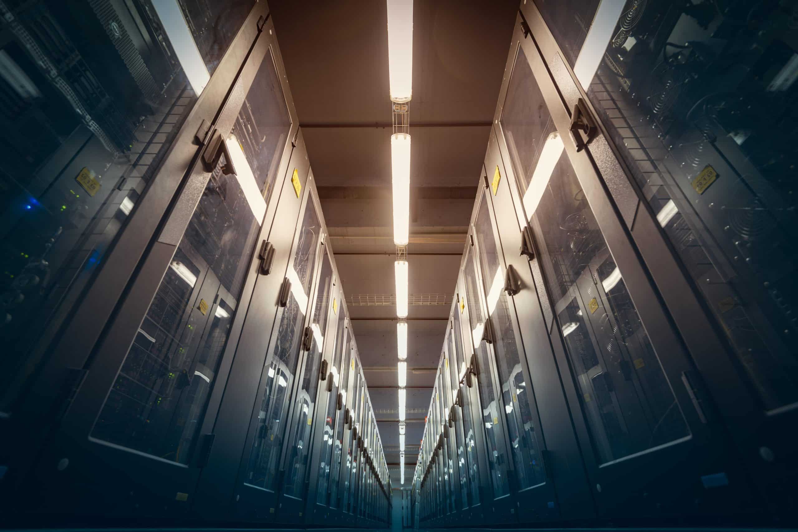 Ein hell erleuchteter Korridor in einem Rechenzentrum, der auf beiden Seiten von hohen, mit Glastüren versehenen Serverschränken gesäumt ist, erstreckt sich unter fluoreszierenden Deckenleuchten in die Ferne.